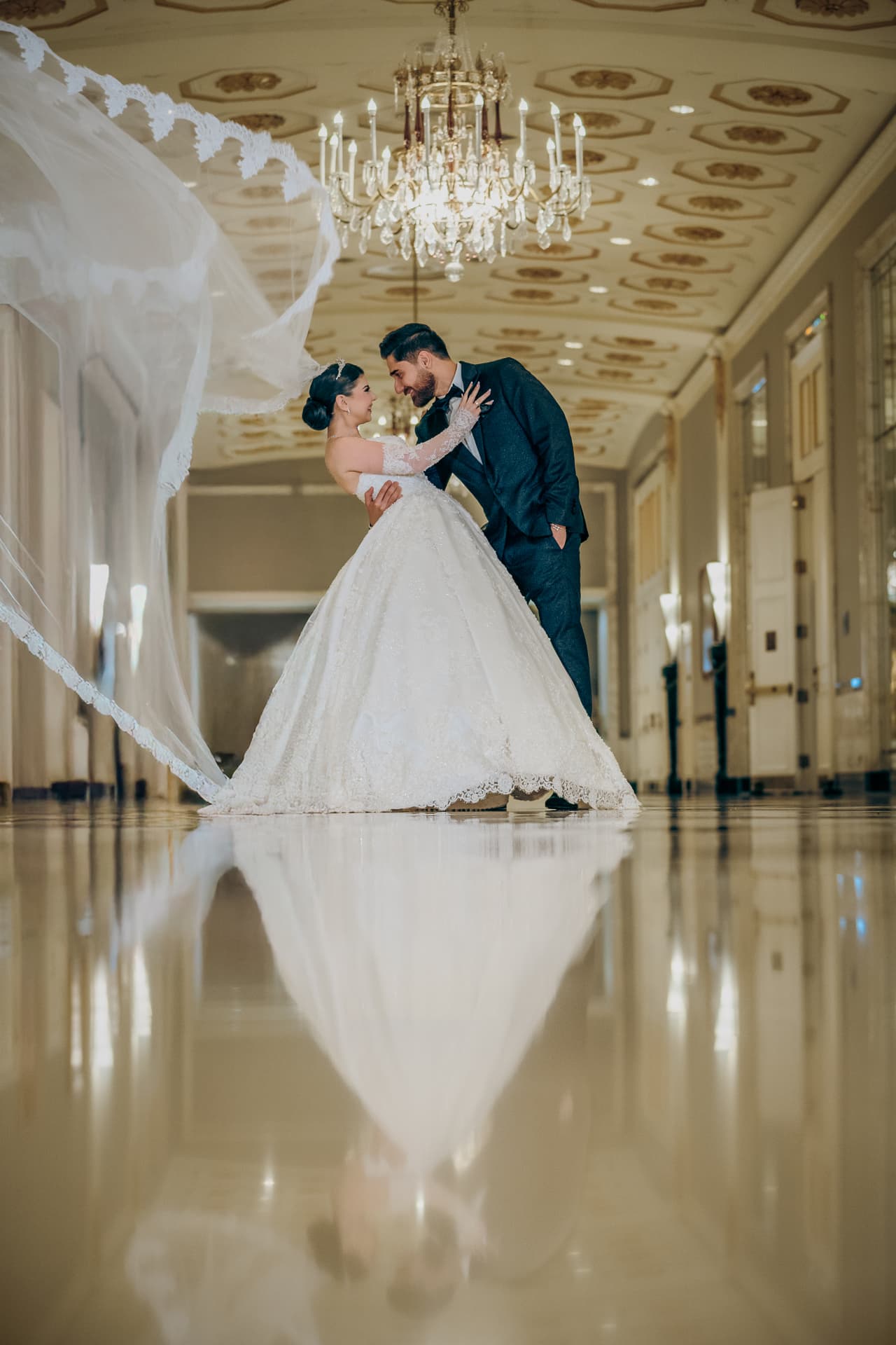 Bride and groom in a grand hallway with a sweeping veil and chandelier reflection