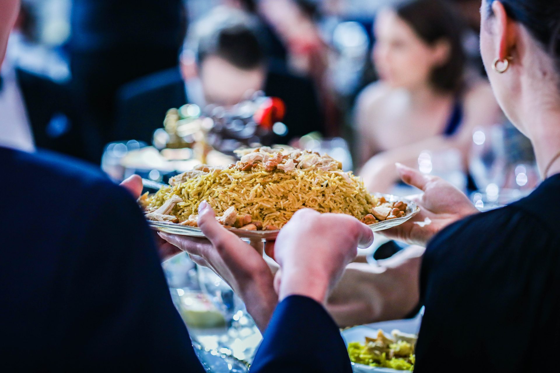 Malak's Kitchen staff serving a platter of rice topped with chicken and toasted nuts to seated guests